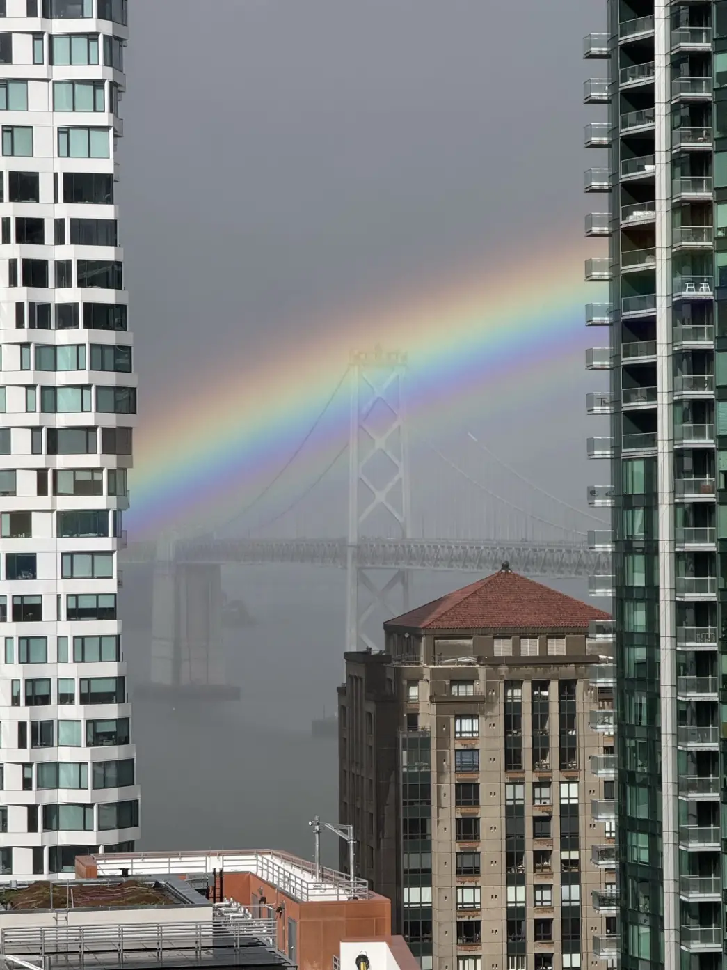 A rainbow arcing between buildings over a bridge on a rainy day.