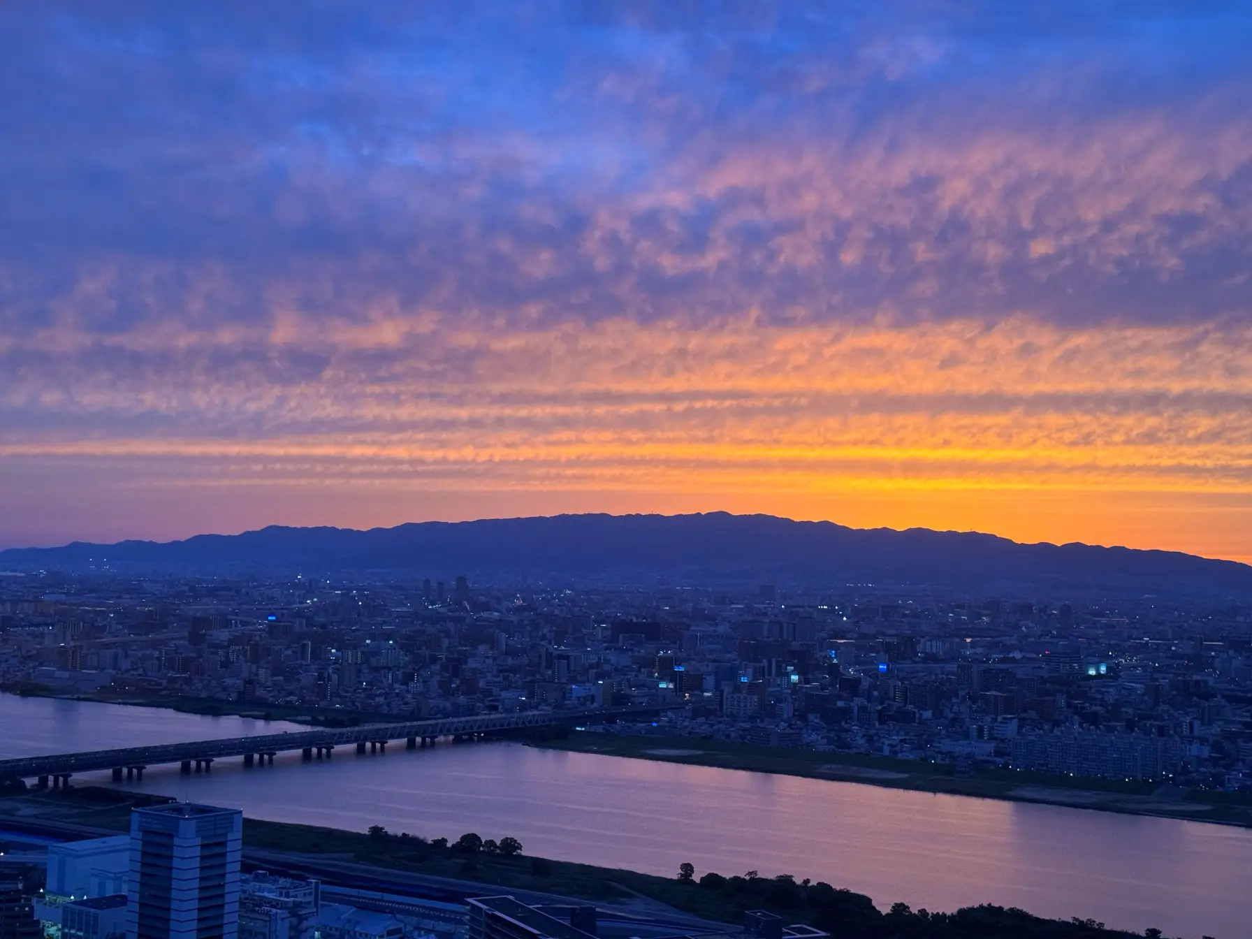 Osaka at sunset from above — a golden-hour gradient spilling over the hills into a blue sky.